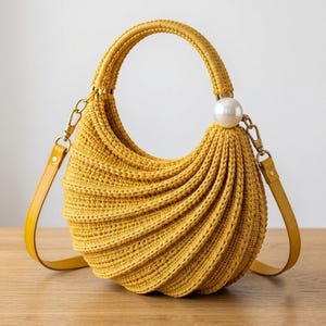 May include: A mustard yellow crescent-shaped handbag crafted from textured crochet material. The bag features a rounded handle, a large white spherical bead accent, and a detachable yellow shoulder strap with gold-toned metal hardware clips.