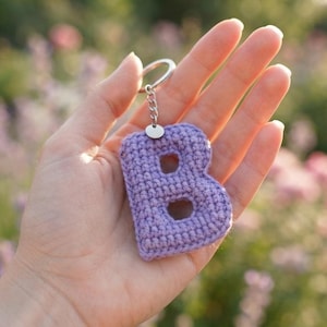 May include: A light purple crochet letter B keychain featuring a textured yarn surface, a small silver-toned metal jump ring, a short chain, and a circular split ring for attachment. The letter is crafted with visible stitches in a soft lavender hue.