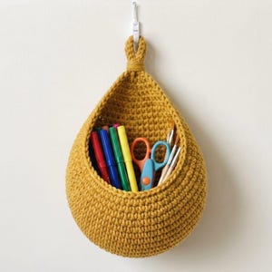 May include: A mustard yellow teardrop-shaped hanging basket made of thick crocheted yarn. The basket is suspended from a white hook and contains a collection of colorful markers, a pair of orange and blue scissors, and several paintbrushes.