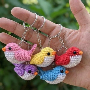 May include: Five small, handcrafted crochet bird keychains in pink, yellow, red, purple, and blue. Each bird features a white belly, a small orange beak, and a black bead eye. The birds are attached to silver-toned metal key rings with chains.
