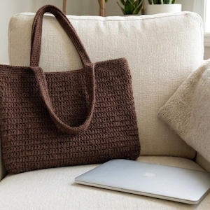 Puede incluir: Un bolso tote rectangular de ganchillo en color marrón oscuro con dos asas a juego, que presenta un patrón de puntos horizontales consistente en todo el cuerpo del bolso, colocado junto a un portátil plateado cerrado.