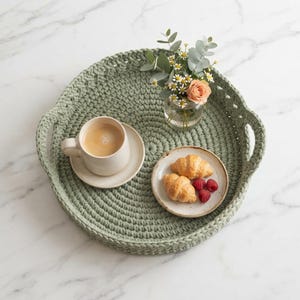 Può includere: Un vassoio rotondo all'uncinetto color verde salvia con manici integrati. Sul vassoio poggiano una tazza in ceramica beige riempita di caffè, un piattino con due croissant dorati e tre lamponi, e un vaso di vetro con un bouquet di fiori.