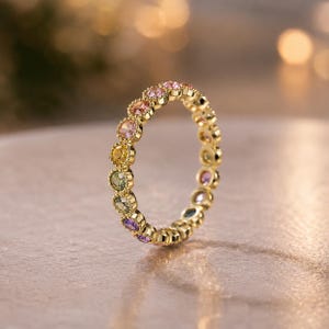 May include: A gold eternity ring featuring a continuous band of round, multi-coloured gemstones including pink, orange, yellow, green, and purple stones. Each gemstone is individually set within a scalloped gold bezel setting along the band.