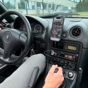 May include: A black car interior featuring a steering wheel with a central logo, a digital clock displaying 12:20, a Pioneer car stereo, and a smartphone mounted on the dashboard displaying a map application connected via a black charging cable.
