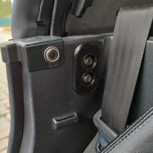 Könnte beinhalten: Eine schwarze Kunststoff-Innenverkleidung für Fahrzeuge mit einer vertikalen Halterung und zwei versenkten Sternschrauben. Die Baugruppe umfasst eine strukturierte Oberfläche, einen freiliegenden Metallschraubenkopf, einen Sicherheitsgurt und einen Schlitz.