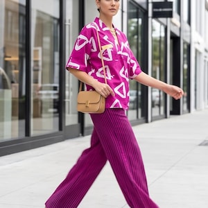 May include: A bright magenta short-sleeved button-down shirt with a white geometric triangle pattern, paired with deep purple pleated wide-leg trousers and a tan leather crossbody bag with a gold clasp, worn with clear heeled sandals.