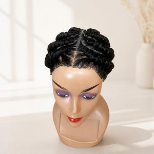 May include: A mannequin head featuring a black wig styled with thick, braided cornrows that run from the front hairline towards the back. The wig has a defined centre part and a smooth, natural-looking hairline, displayed on a smooth, light-toned base.
