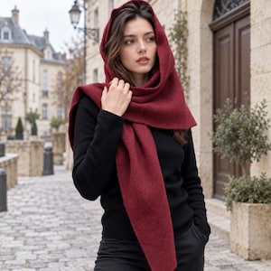 May include: A deep burgundy, textured wool-blend scarf draped over the head and shoulders, worn over a solid black long-sleeved shirt. The scarf features a thick, soft-knit fabric with a visible weave, providing full coverage around the neck.