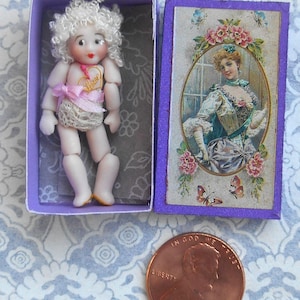 May include: A miniature doll with curly white hair, red lips, and a gold heart charm, resting in a purple box next to its lid featuring a vintage illustration of a woman. A copper penny is placed nearby for scale, showing the small size.