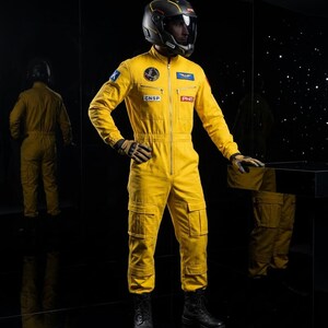 May include: A bright yellow flight suit featuring a front zipper, multiple cargo pockets on the legs, and various patches including CNSP and PHM. The suit is worn with a black helmet, black gloves, and black boots, creating a cohesive aviation look.