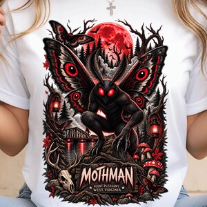 Può includere: Una t-shirt bianca con una stampa grafica di una creatura Mothman nera con grandi occhi rossi e ali, su sfondo di luna rossa, foresta e ponte. Il testo recita MOTHMAN POINT PLEASANT WEST VIRGINIA, accompagnato da un teschio di cervo e funghi.