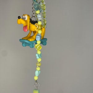 Puede incluir: Un llavero con una figura de perro de dibujos animados amarilla sobre un monopatín azul cerceta, unido a un clip de plástico amarillo. El dije incluye una cadena de cuentas translúcidas amarillas y azul claro con formas de corazones y estrellas.