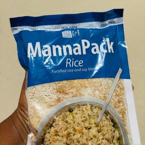 May include: A blue and white plastic pouch of MannaPack Rice, a fortified rice and soy blend. The packaging features the text MannaPack Rice, Feed My Starving Children, and Seal Area, with a graphic of a bowl of cooked rice and a spoon.
