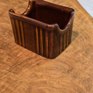 May include: A rectangular brown ceramic holder with a glossy finish and vertical ribbed detailing along the exterior walls. The container features a U-shaped cutout on the front and back panels, creating a low-profile opening for easy access.