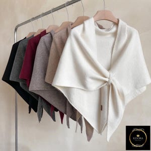 May include: A row of six triangular knit shawls hanging on a metal rack, featuring colors including white, beige, gray, burgundy, dark gray, and black. Each shawl has a knotted front detail and a small brown tag with the text KESORA.