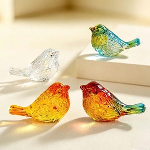 May include: Four small glass bird figurines. One is clear, one is blue and green, one is yellow and orange, and one is red and orange. Each bird features molded feather details, a small beak, and a rounded body shape, arranged in a group.