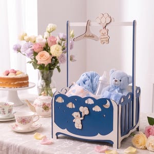May include: A blue and white wooden cradle-shaped storage basket featuring a cutout bear holding balloons. The basket includes a hanging wooden hanger with the name Arabella and a bear ornament, containing a light blue teddy bear and baby items.