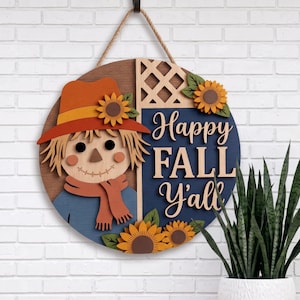 May include: A circular wooden wall sign featuring a scarecrow wearing an orange hat and scarf, accented with sunflowers and a lattice pattern. The sign displays the text "Happy FALL Y'all" in white lettering against a dark blue background.