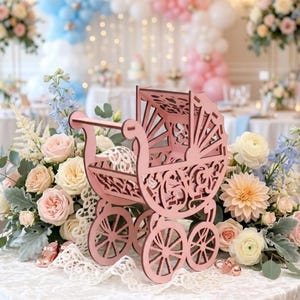 May include: A dusty pink laser-cut wooden baby carriage centerpiece featuring intricate floral and scrollwork patterns on the sides and hood, supported by four spoked wheels, displayed on a white lace-covered table with floral arrangements.