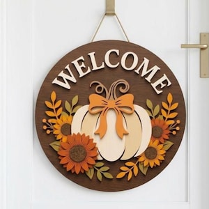May include: A circular dark brown wooden sign featuring the word WELCOME in raised white letters at the top. The center displays a light-colored pumpkin with an orange bow, surrounded by layered orange and yellow sunflowers and green leaves.