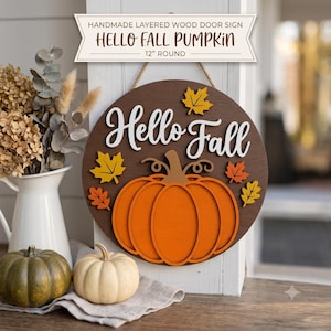 May include: A 12-inch round layered wood door sign featuring a dark brown background with the white text Hello Fall. The sign includes a large orange pumpkin cutout and several small yellow and orange maple leaf accents, suspended by a jute rope.