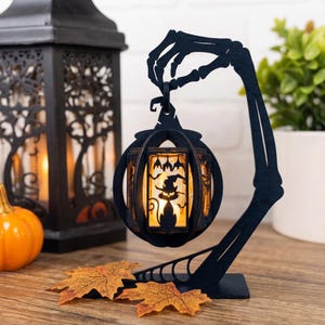 May include: A black wooden decorative stand shaped like a skeleton hand holding a circular lantern. The lantern features a cutout silhouette of a cat wearing a witch hat and bats, illuminated from within by a warm glowing light.