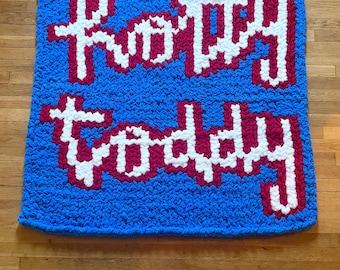 May include: A chunky knit blanket in bright blue featuring the text Hotty Toddy in white yarn with a dark red outline. The letters are crafted in a blocky, pixelated style using thick, soft yarn loops throughout the entire rectangular piece.