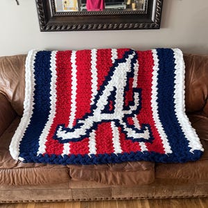 May include: A chunky knit throw blanket featuring a red, white, and navy blue striped pattern. The center of the blanket displays a large, stylized white letter A with a navy blue outline, mimicking a professional baseball team logo design.