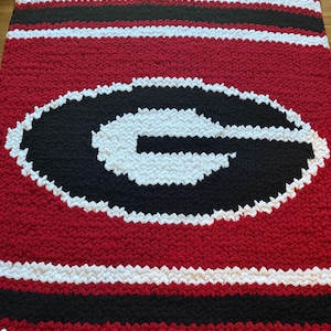 May include: A chunky knit blanket featuring a large black letter G outlined in white on a red background. The blanket has horizontal stripes of red, white, and black at the top and bottom edges, created with thick, soft yarn in a textured pattern.