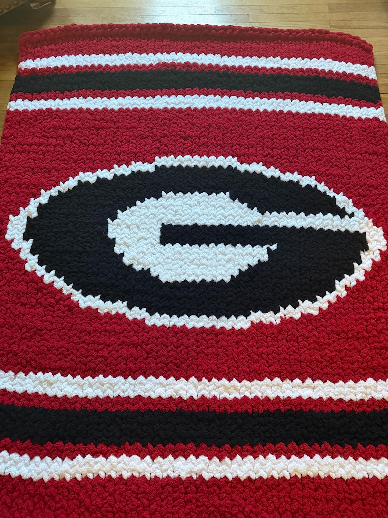 May include: A chunky knit blanket featuring a large black letter G outlined in white on a red background. The blanket has horizontal stripes of red, white, and black at the top and bottom edges, created with thick, soft yarn in a textured pattern.