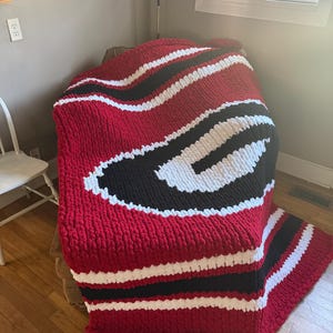 May include: A chunky hand-knitted blanket featuring a large letter G logo in white and black, set against a deep red background. The throw is crafted from thick, soft yarn with a prominent textured stitch pattern and horizontal stripes in red, white, and black.