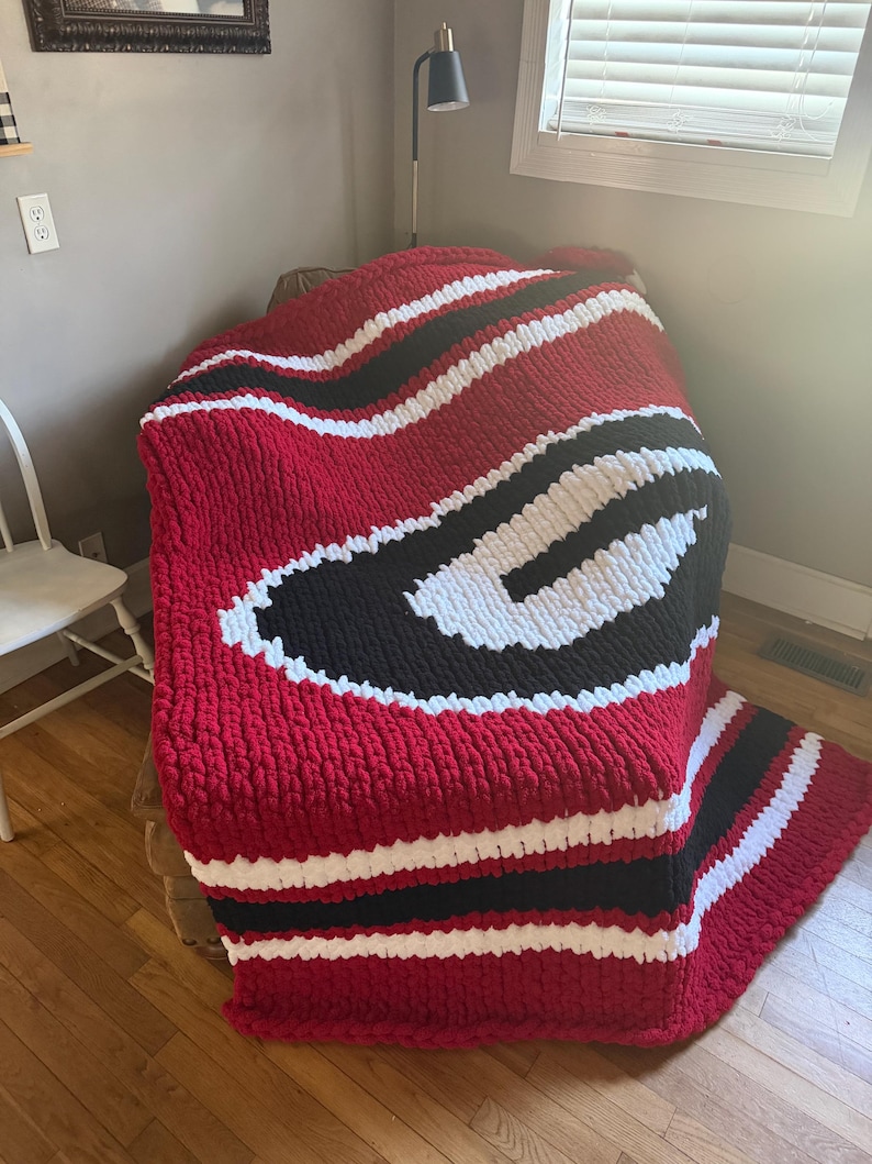 May include: A chunky hand-knitted blanket featuring a large letter G logo in white and black, set against a deep red background. The throw is crafted from thick, soft yarn with a prominent textured stitch pattern and horizontal stripes in red, white, and black.