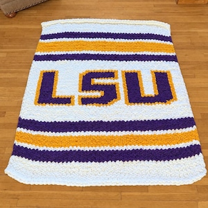May include: A rectangular hand-knitted blanket featuring a chunky white, purple, and gold striped pattern. The centre of the blanket displays the letters LSU in large, bold purple block characters outlined in gold yarn against a white background.