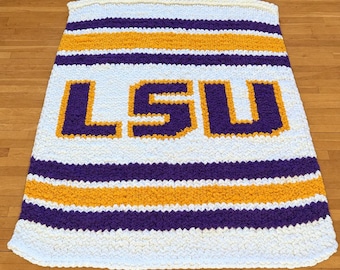 May include: A rectangular hand-knitted blanket featuring a chunky white, purple, and gold striped pattern. The center of the blanket displays the letters LSU in large, bold purple block characters outlined in gold yarn against a white background.