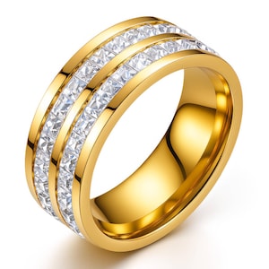 May include: A gold-toned band ring featuring two parallel channels filled with clear, square-cut stones. The ring has a polished, reflective surface and a wide, smooth profile, showcasing a continuous row of sparkling stones set within the metal.