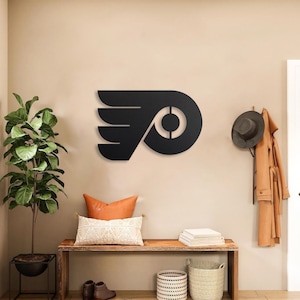 Op de afbeelding: Een matzwart metalen wanddecoratie met het logo van de Philadelphia Flyers, gekenmerkt door een gestileerde gevleugelde letter P met een cirkelvormig midden, centraal gemonteerd op een lichtbeige muur boven een houten bank.