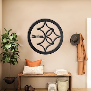 Op de afbeelding: Een cirkelvormig wandkunstobject van zwart metaal met het logo van de Pittsburgh Steelers, waarbij het woord Steelers aan de linkerkant is geïntegreerd. Het ontwerp bestaat uit een dikke buitenring die drie vierpuntige sterren omsluit.