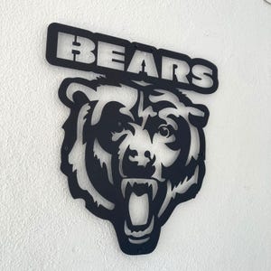 Può includere: Un'insegna da parete in metallo nero raffigurante una testa di orso stilizzata con la bocca aperta e i denti in mostra. Sopra la testa dell'orso, un banner rettangolare in metallo mostra la parola BEARS in lettere maiuscole in grassetto.