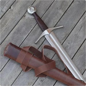 Può includere: Una spada in acciaio con impugnatura rivestita in pelle marrone scuro, pomolo circolare argentato e guardia dritta. La spada è accompagnata da un fodero in pelle marrone scuro abbinato con cinghie in pelle integrate.