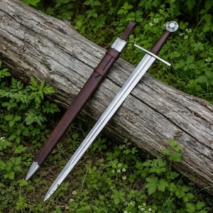 Op de afbeelding: Een stalen zwaard met een recht, tweesnijdend lemmet, een eenvoudige pareerstang en een donkerbruin, met leer omwikkeld handvat met een ronde pommel. Ernaast ligt een bijpassende donkerbruine lederen schede met een zilverkleurige punt en mond.