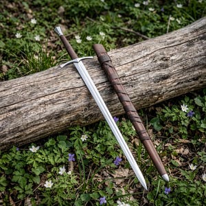 Pode incluir: Uma espada de aço em tom prateado com punho revestido em couro castanho escuro e uma bainha de couro castanho escuro a condizer com um design de tiras cruzadas. A espada tem uma guarda reta e uma lâmina pontiaguda, pousada num tronco de madeira.