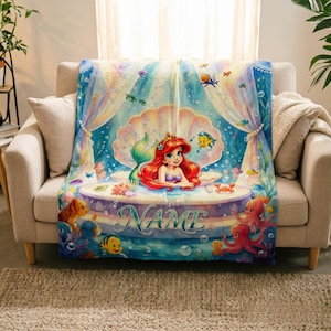 May include: A soft fleece blanket featuring a colourful underwater scene with a red-haired mermaid character sitting inside a large seashell. The design includes various sea creatures, bubbles, and the word NAME printed in a decorative script font.