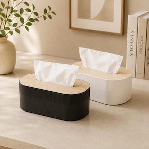 May include: Two oval tissue box covers with light wood grain lids and ribbed sides. One cover is matte black and the other is matte white. Each box features a central rectangular opening with a white tissue pulled through the top surface.
