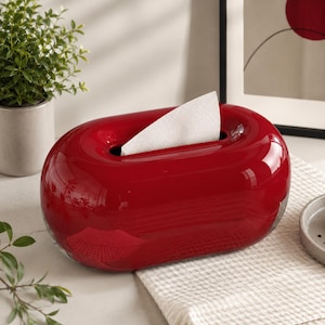 Könnte beinhalten: Ein glänzender, oval geformter Taschentuchbox-Behälter in leuchtendem Rot. Das Objekt hat ein glattes, abgerundetes Design mit einer rechteckigen Öffnung in der Mitte, aus der ein einzelnes weißes Papiertaschentuch teilweise herausgezogen ist.