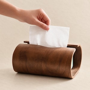 Puede incluir: Un soporte para caja de pañuelos de madera marrón oscuro curvada con un acabado liso y pulido. La estructura rectangular presenta una ranura superior abierta por donde se extrae hacia arriba un pañuelo de papel blanco del compartimento interior.