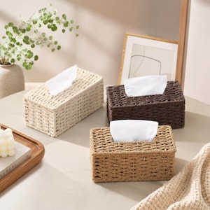 May include: Three rectangular tissue box covers made of woven material in cream, dark brown, and light tan colors. Each cover features an open top slot with a white tissue protruding from the center, showcasing a textured, grid-like weave pattern.