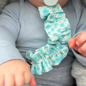 May include: A pacifier clip featuring a ruffled fabric strap with a light blue and white floral pattern. The clip is attached to a white pacifier with a circular center, and the fabric strap is gathered with a ruffled edge along its length.