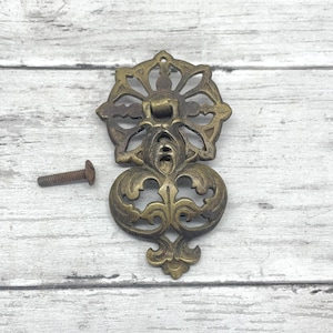 May include: An antique brass drawer pull with a decorative floral design and a matching screw. The pull has an ornate, symmetrical shape with a central flower-like element and scrolling details. The hardware is likely for a cabinet or drawer.