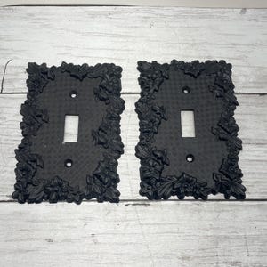 May include: Two black, decorative light switch covers. Each cover has a rectangular shape with a floral design around the edges. The covers have a single rectangular opening for the switch and two screw holes.