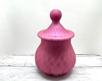 May include: A pink, lidded jar with a textured surface. The jar has a bulbous body and a lid with a rounded finial. The jar is made of a solid pink material and sits on a white wooden surface.